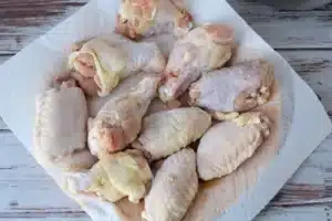 A collection of raw chicken wings and drumsticks are laid out on a white paper towel on a rustic, white wooden table. The fresh chicken pieces are ready for preparation or cooking, and would be perfect as air fryer chicken wings when seasoned with a touch of baking powder.