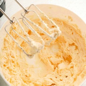 A mixing bowl filled with creamy, light yellow butter awaits. An electric mixer with two beaters, coated in the same velvety mixture for a luscious brownie frosting, rests above the bowl on a pristine white countertop.