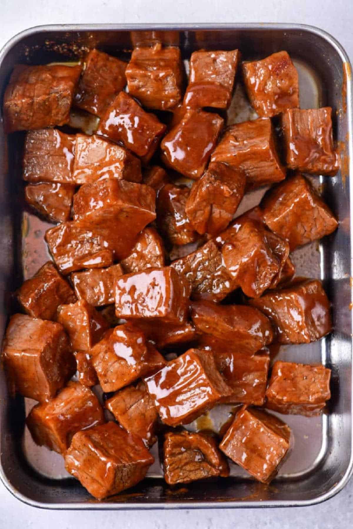 Cubed pieces of marinated beef, inspired by Chuck Roast Burnt Ends, rest in a metal tray coated with a glossy, reddish-brown sauce.