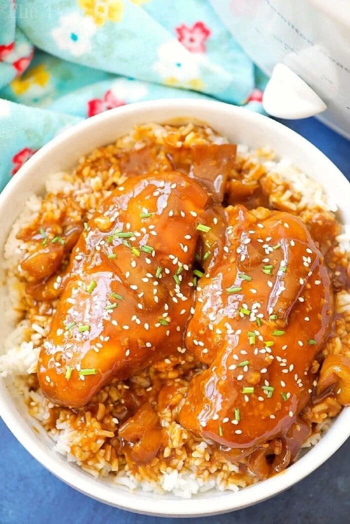 This easy crockpot sticky teriyaki chicken with a kick of heat recipe is amazing and a healthy meal too! Sweet with just a pinch of Sriracha makes it great! Bowl of rice topped with crockpot teriyaki chicken, chopped herbs, sesame seeds, and a savory brown sauce.
