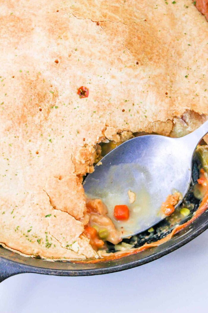 A spoon scooping vegetables and sauce from a skillet with a golden-brown pie crust on top, revealing the hearty filling of a Leftover Steak Pot Pie.
