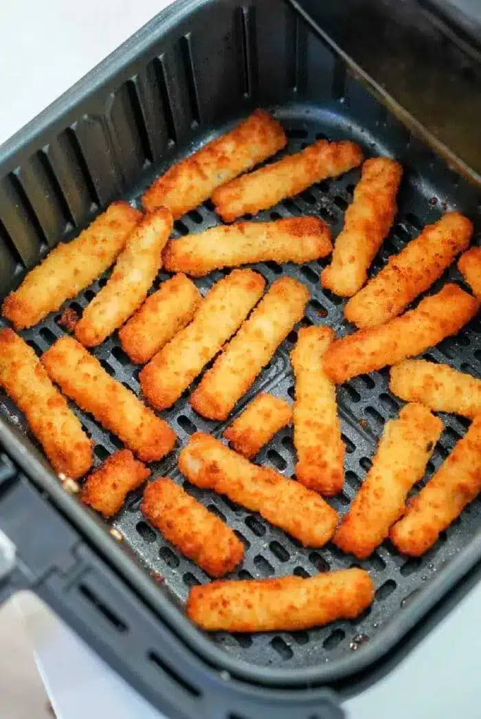 Several air fryer fish sticks are arranged in a single layer inside an open air fryer basket. The fish sticks are golden brown and appear to be evenly cooked. The air fryer basket is black and has a perforated bottom.