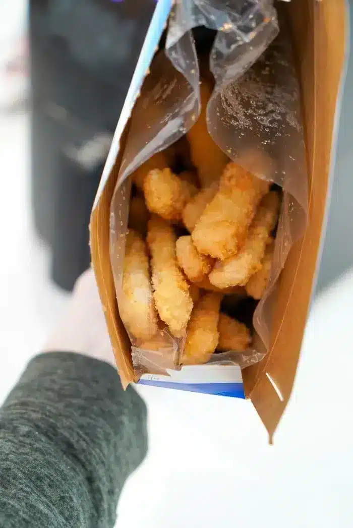 An open cardboard box filled with frozen fish sticks can be seen, ideal for making air fryer fish sticks. The fish sticks are partially wrapped in clear plastic inside the box. 