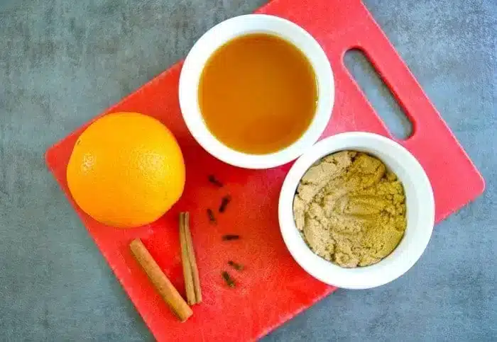 A red cutting board holds an orange, a small bowl of brown sugar, a small bowl of liquid, cinnamon sticks, and cloves on a gray surface—perfect ingredients for crafting crockpot hot apple cider.