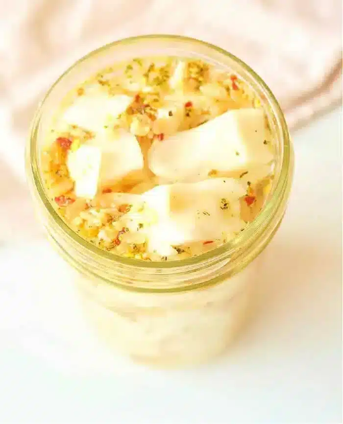 A jar filled with marinated cheese cubes, topped with herbs and seasoning, sits alongside a vibrant batch of quick-pickled cabbage. The scene is captured from a high angle on a light-colored surface, creating a delightful culinary tableau.