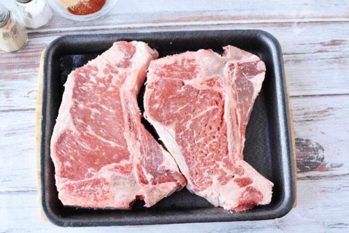 Two raw pork chops in a black tray on a light wooden surface, with spices visible in the corner—perfect for preparing dishes like Leftover Steak Pot Pie or your favorite hearty meal.