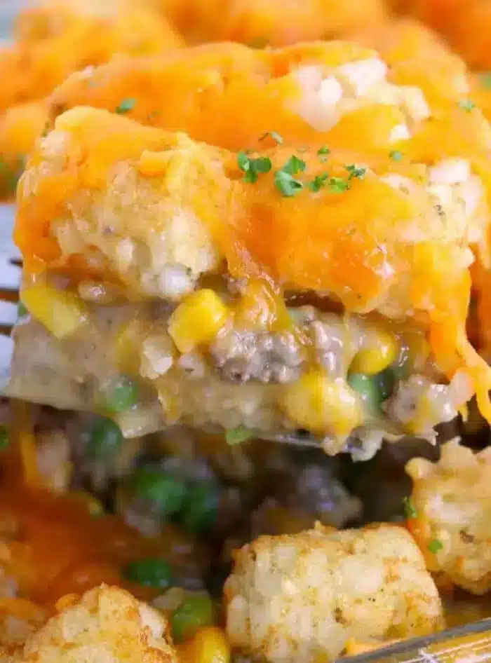 A fork holds a piece of potato tot casserole with ground beef, revealing layers of savory ground beef, mixed vegetables, and melted cheddar cheese. This dish delights with corn, peas, and cheese-topped tater tots, all garnished with a sprinkle of parsley.