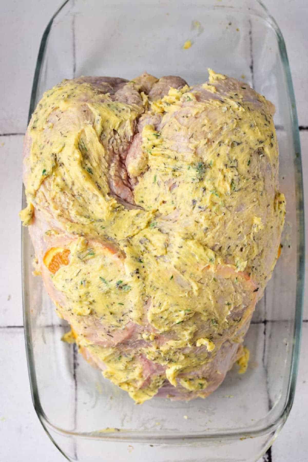 Raw Turkey Crown coated with a yellow herb butter mixture in a glass baking dish.
