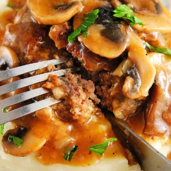 A fork cuts into a Salisbury steak with mushroom gravy atop mashed potatoes, reminiscent of delightful ground venison recipes.