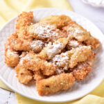 A plate of crispy banana tempura dusted with powdered sugar sits elegantly on a white plate atop a yellow cloth.