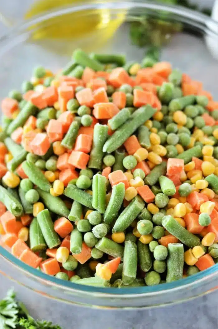 A glass bowl brimming with frozen mixed vegetables—green beans, peas, carrots, and corn—awaits the magic of roasting. The gray countertop sets a subtle stage with a hint of parsley for garnish.