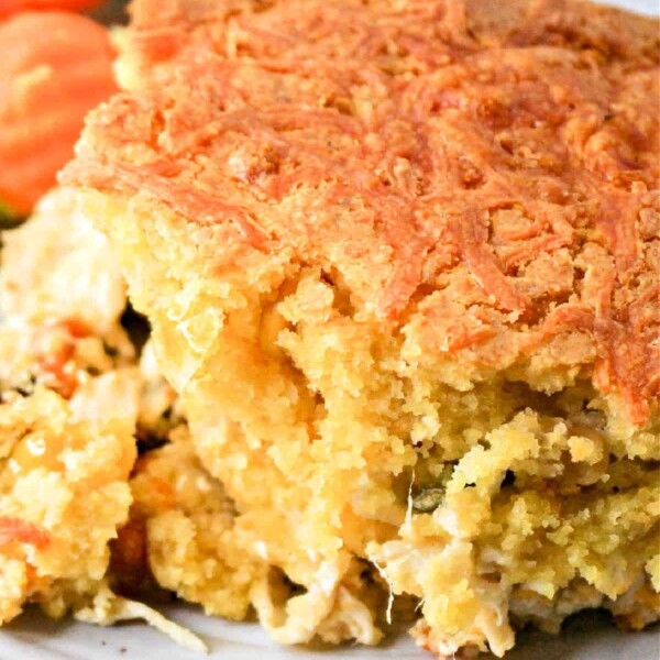 Close-up of a portion of cornbread casserole with a golden crust, served alongside fresh broccoli and sliced carrots on a white plate.