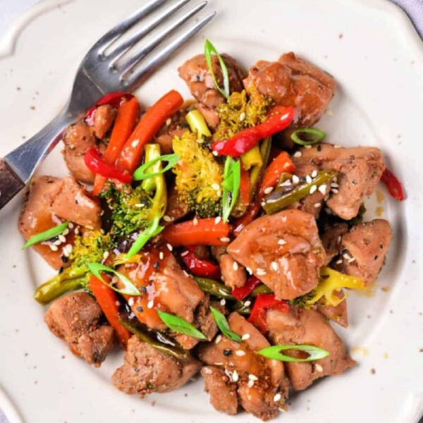 Savor the delicious Chicken Thigh Stir Fry featuring tender chicken, broccoli, bell peppers, and sesame seeds artfully arranged on a white plate with a fork.