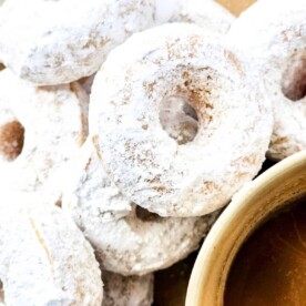 A close-up of several powdered sugar donuts stacked on a plate, with a cup filled with a rich, brown liquid, likely coffee or tea.