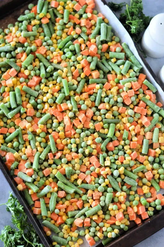 A baking sheet brimming with roasting frozen vegetables, including green beans, corn, peas, and diced carrots. Fresh parsley peeks out in the background.