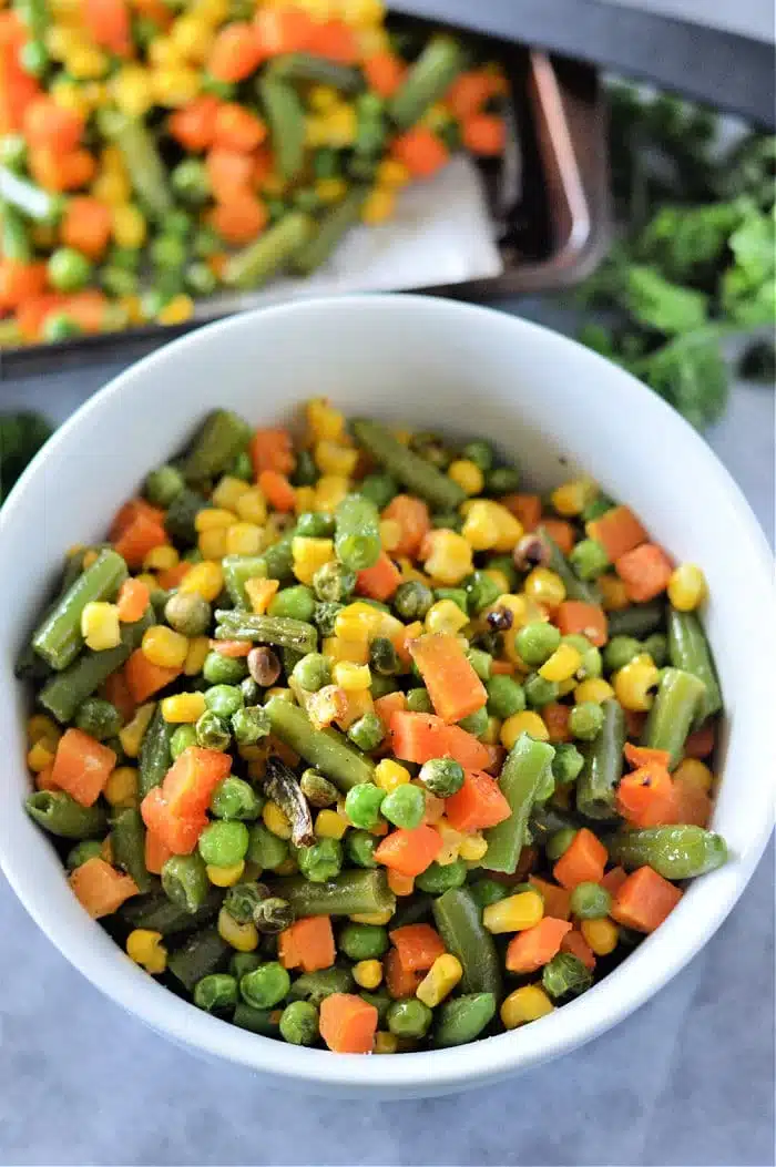 A bowl filled with roasted mixed vegetables, including green beans, corn, peas, and diced carrots. The vibrant colors suggest they were perfectly cooked from frozen. In the background, more vegetables await roasting on a tray.