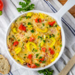 A delicious baked dish of Rotel Sausage Dip with melted cheese, peppers, and herbs in a white pan on a cloth, perfectly paired with a fresh baguette nearby.