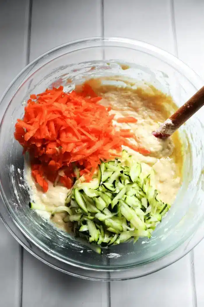 A glass bowl contains batter mixed with grated carrots and zucchini, perfect for making delicious zucchini carrot muffins. A wooden spoon is partially submerged in the mixture, resting on a white countertop.