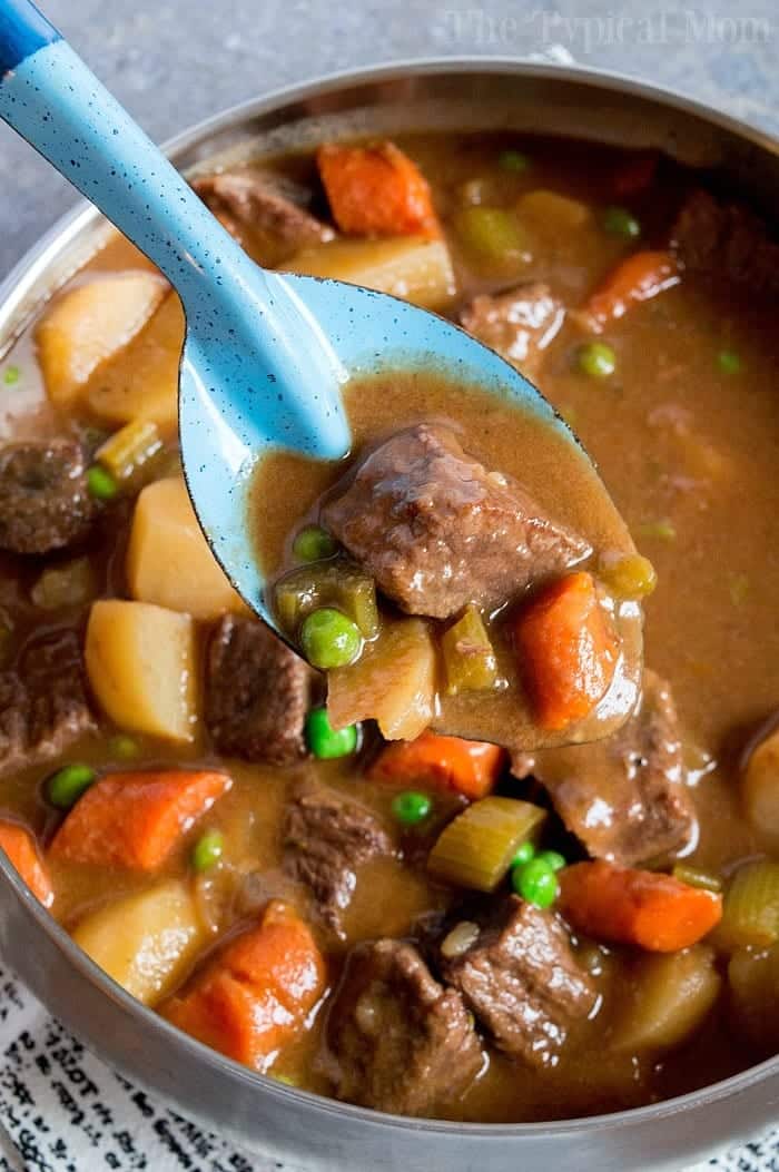 A ladle serving hearty beef stew with carrots, peas, and potatoes from a rustic Dutch oven.