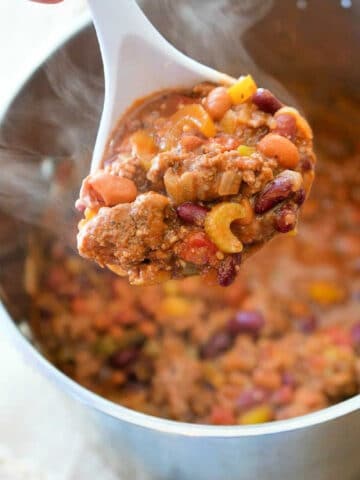 A white ladle holds a hearty blend reminiscent of an easy Bloody Mary chili recipe, featuring kidney beans, ground meat, and diced vegetables over a steaming pot filled with the same savory mixture.