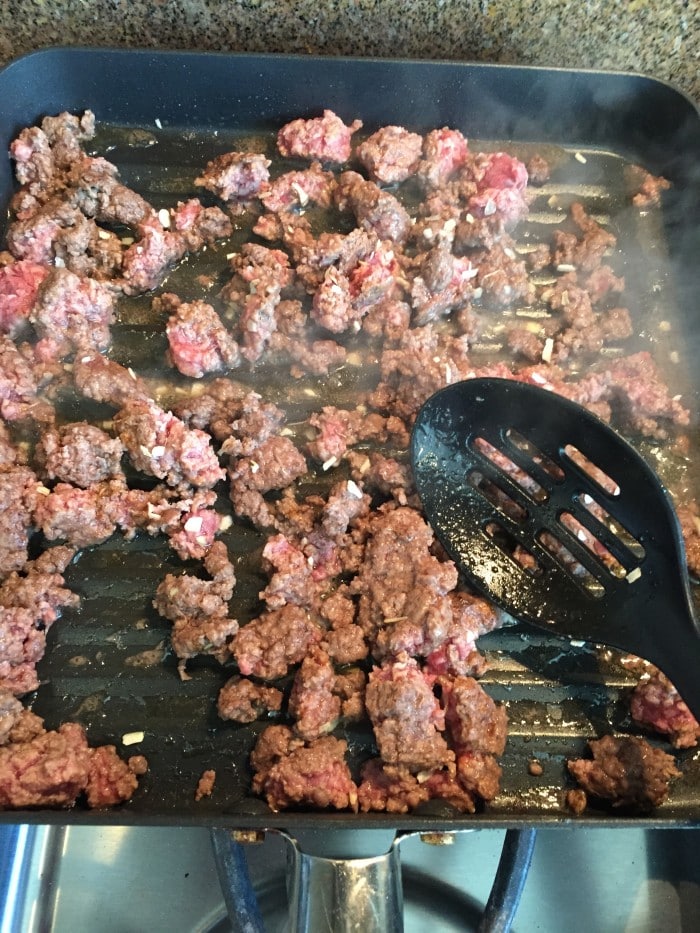 Ground beef is sizzling in a grill pan on the stovetop, perfect for a mild chili recipe. A black slotted spatula rests nearby as steam rises from the browning meat. The pan's grill lines sear the beef while bits of onion add flavor, hinting at delicious possibilities ahead.