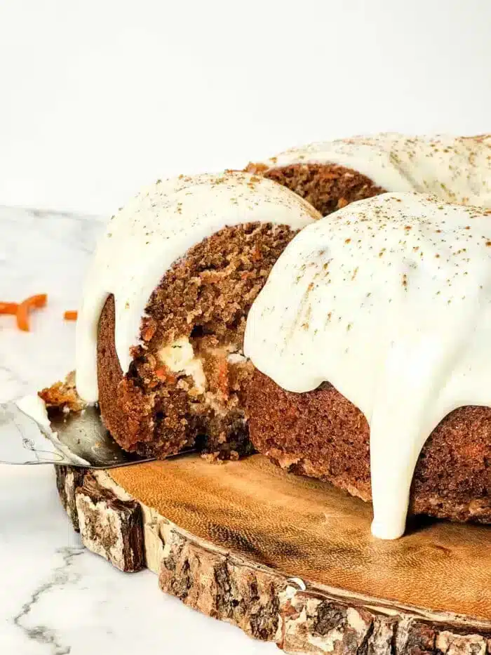 A slice of sumptuous carrot Bundt cake with cream cheese frosting is being served. The cake rests elegantly on a wooden platter. The thick frosting drapes the top, with grated carrots adding a rustic touch nearby on the marble surface.