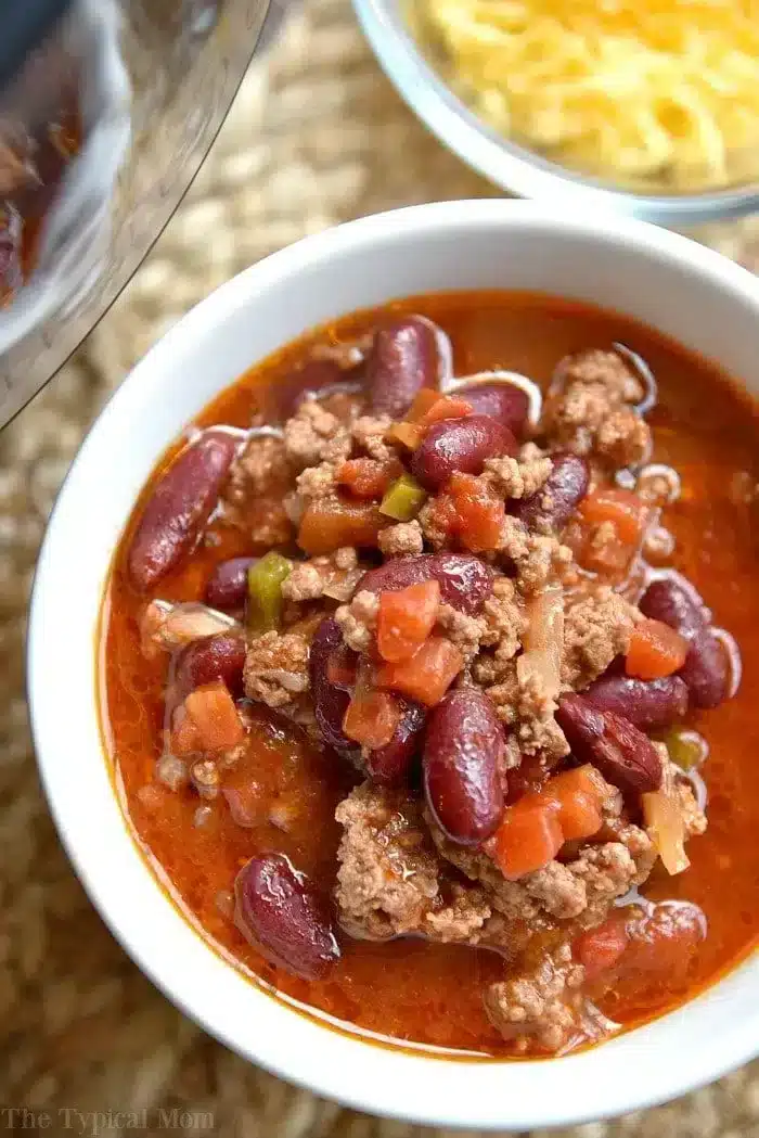 A bowl of chili featuring ground meat, kidney beans, and diced vegetables in a tomato-based sauce. Reminiscent of an easy bloody mary chili recipe, a bowl of shredded cheese is blurred in the background. The table is covered with a textured, woven mat.