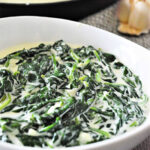 A white bowl filled with creamy spinach, showcasing delicious creamed spinach, with a pan and garlic in the background.