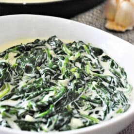 A white bowl filled with creamy spinach, showcasing delicious creamed spinach, with a pan and garlic in the background.