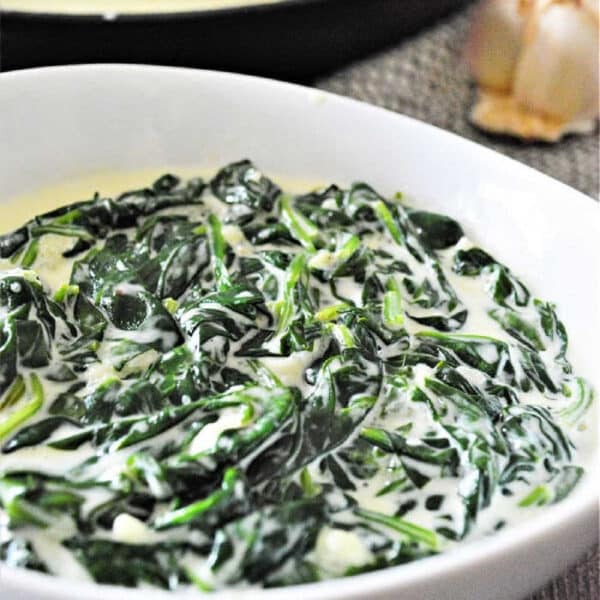 A white bowl filled with creamy spinach, showcasing delicious creamed spinach, with a pan and garlic in the background.