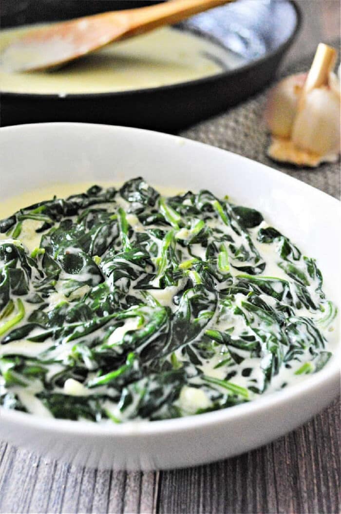 A white bowl filled with creamy spinach, showcasing delicious creamed spinach, with a pan and garlic in the background.