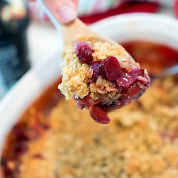 A hand holds a wooden spoon with a scoop of cherry crisp recipe above a baking dish.