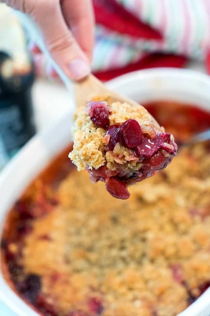 A hand holds a wooden spoon with a scoop of cherry crisp recipe above a baking dish.