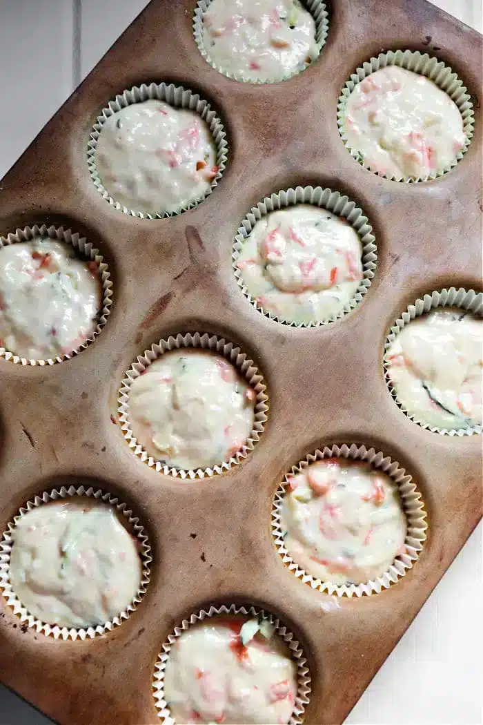 A muffin tin holds eleven cupcake liners filled with zucchini carrot muffins batter, showcasing pieces of red and green ingredients. The tin rests on a white surface.