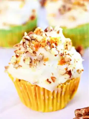 A mini zucchini carrot cupcake with white frosting and chopped nuts sits in a yellow liner with white polka dots. Blurred cupcakes are visible in the background.