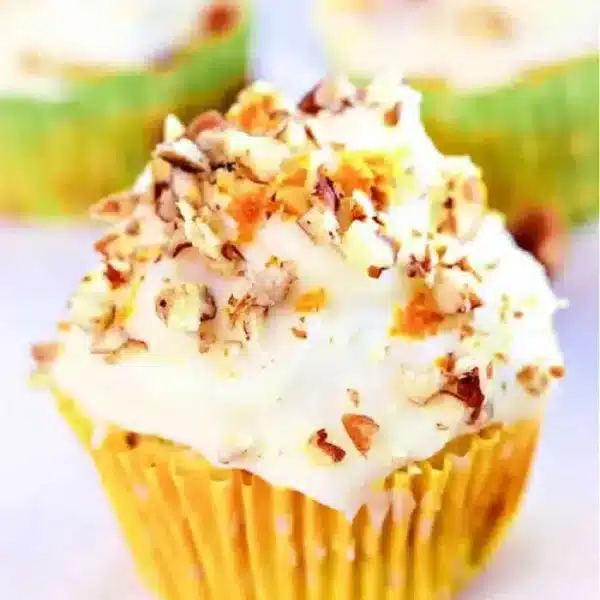 A mini zucchini carrot cupcake with white frosting and chopped nuts sits in a yellow liner with white polka dots. Blurred cupcakes are visible in the background.