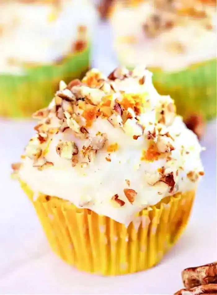 A cupcake with white frosting is topped with chopped nuts, nestled in a yellow polka dot wrapper. In the background, zucchini carrot muffins offer a colorful blend of flavors and blur into the scene.