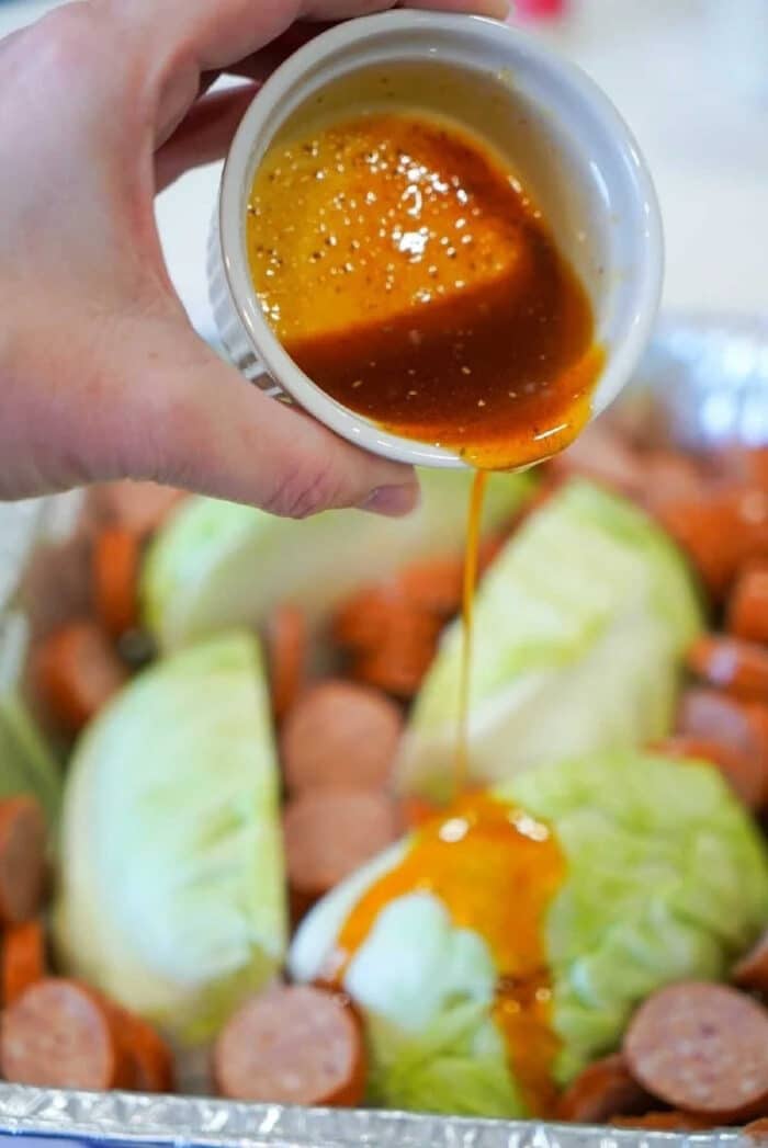 A hand pours sauce from a small bowl over smoked sausage and cabbage wedges in a foil-lined pan.