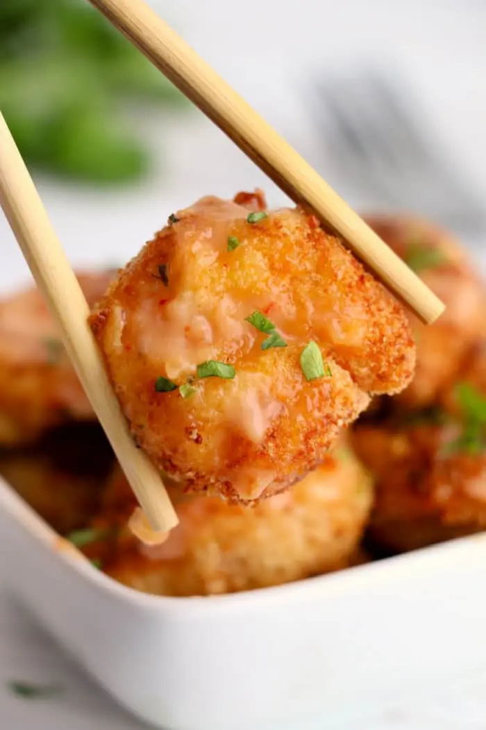 A close-up of chopsticks holding a piece of breaded and fried tofu, garnished with chopped herbs and a light sauce that gives it an almost bang bang shrimp vibe. In the background, there is a square white bowl filled with more air-fried pieces of the same delicious breaded tofu.