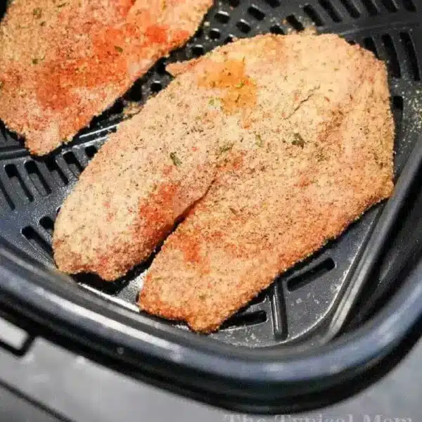 Two breaded tilapia fillets are placed inside an air fryer basket. The fillets are seasoned with a mixture of spices and breadcrumbs, ready for cooking. The air fryer setup indicates the beginning of the cooking process.