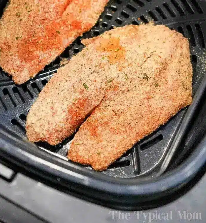 Two breaded tilapia fillets are placed inside an air fryer basket. The fillets are seasoned with a mixture of spices and breadcrumbs, ready for cooking. The air fryer setup indicates the beginning of the cooking process.