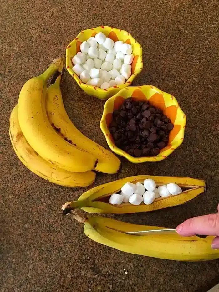 A bunch of bananas sits on a countertop beside two bowls: one filled with mini marshmallows and the other with chocolate chips. A hand cuts open a banana, turning it into a delightful banana s'mores treat by stuffing it with mini marshmallows.