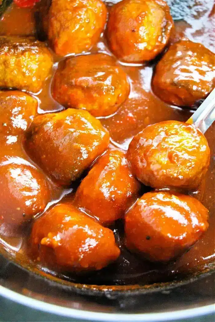 A close-up of delightful meatballs, perfect as a crockpot appetizer, coated in a glossy sauce and placed in a pan. The thick, rich sauce covers each meatball evenly. A utensil is partially visible on the right side, ready to serve.