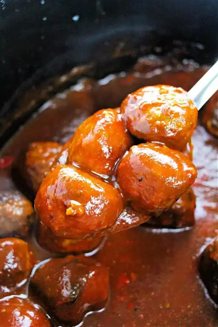 A spoon lifts several glazed meatballs, perfectly cooked as a crockpot appetizer, from a pot filled with thick, brown sauce. The glossy surface of the sauce clings to the meatballs, suggesting they are beautifully marinated and ready to delight at any gathering.