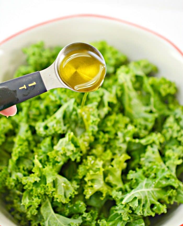 A tablespoon of olive oil is being drizzled over fresh kale in a white bowl, setting the stage for delicious baked kale chips.