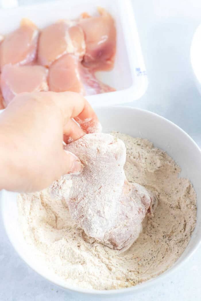 Hand coating raw chicken breast in seasoned flour in a white bowl, with more chicken pieces—including air fryer fried chicken thighs—waiting in a tray nearby.