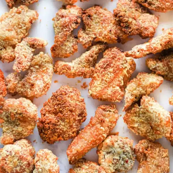 A collection of crispy, golden-brown popcorn chicken pieces arranged on a white surface. The air fryer ensures each piece achieves that textured, crunchy coating, with the pieces varying in size and shape.