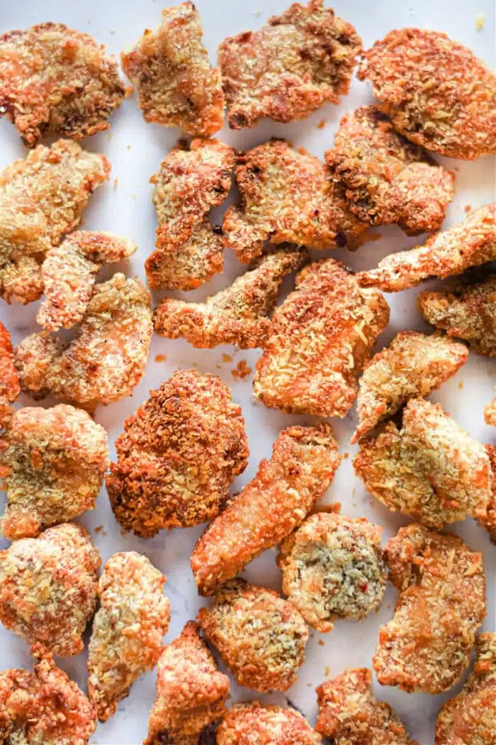 A collection of crispy, golden-brown popcorn chicken pieces arranged on a white surface. The air fryer ensures each piece achieves that textured, crunchy coating, with the pieces varying in size and shape.