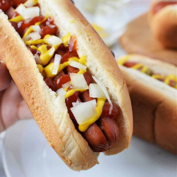 Close-up of a hand holding a hot dog with mustard, ketchup, and chopped onions on a plate, fresh from being cooked to perfection in the oven.