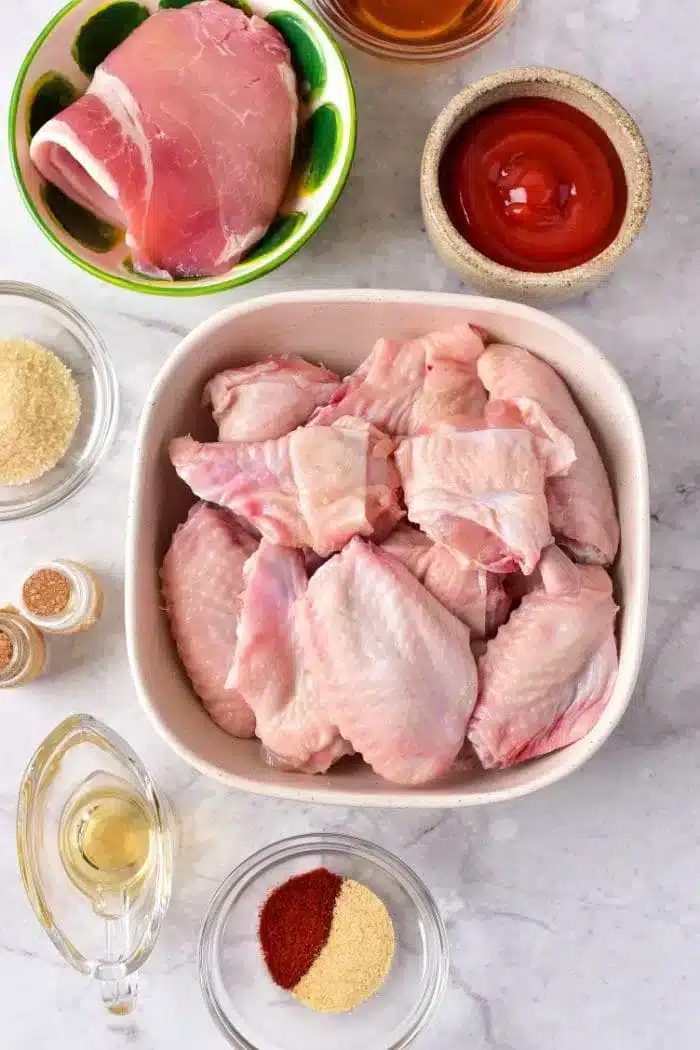 A top-down view of raw chicken wings in a white dish, a bowl of raw boneless chicken thigh, a bowl of red sauce, a small bowl of brown sugar, a small bowl of spices, and a clear jug of oil.